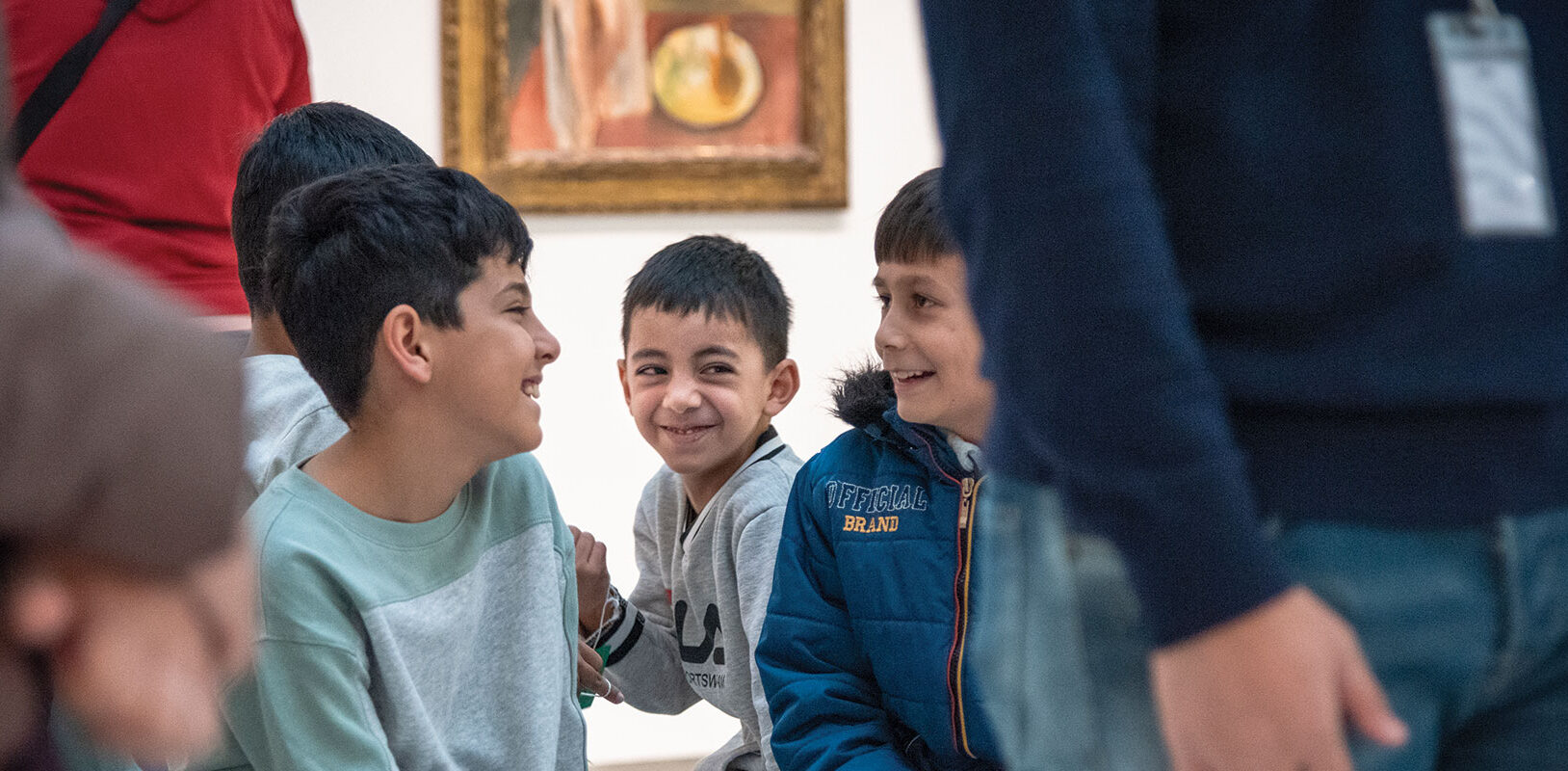 A group of smiling children sit together in an art gallery, sharing laughter and joy, with paintings visible in the background.