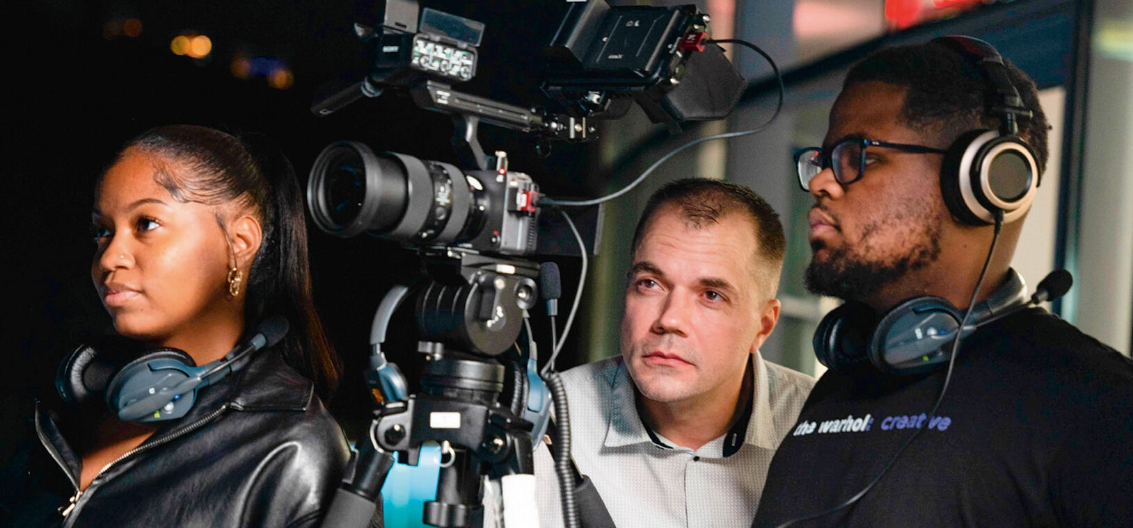 A diverse team of three people reviews footage on set. A woman with headphones, a man focused on the camera, and another wearing a headset.