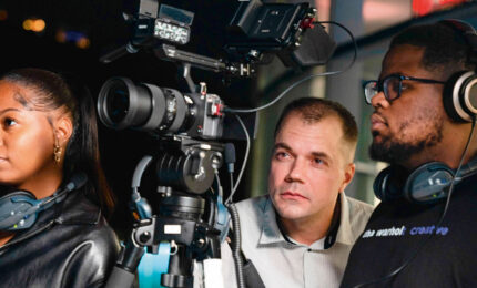A diverse team of three people reviews footage on set. A woman with headphones, a man focused on the camera, and another wearing a headset.