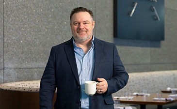 A smiling man in a dark blazer holds a coffee cup, standing in a modern café with a textured wall in the background.