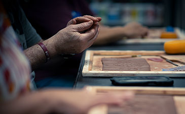 Close-up of hands crafting with clay at a workshop, with textured brown clay pieces and tools on the work surface.