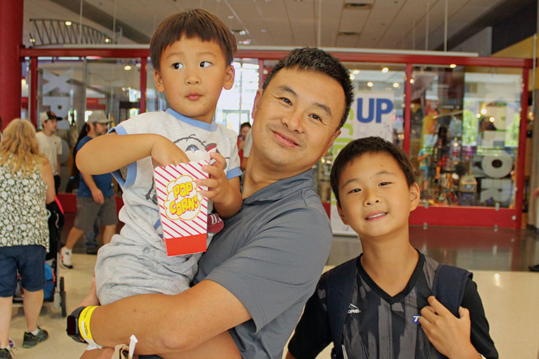 A smiling man holds a young boy with popcorn while another boy stands beside them, all in a lively indoor museum setting.