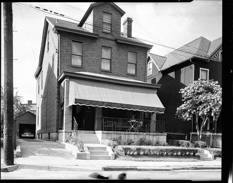 A historic three-story brick house with a front porch and awning, surrounded by gardens and nearby trees, in black and white.