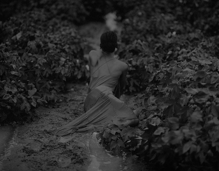 A person in a flowing dress sits in a muddy path surrounded by lush green foliage, evoking a serene yet mysterious atmosphere.